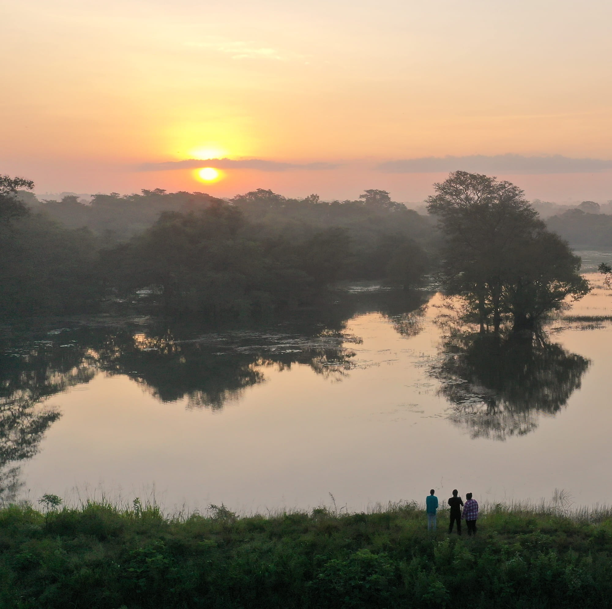 Three people standing near a lake. Sun is setting in the background.